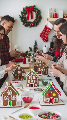 group of people standing around a table with gingerbread houses on it and bowls of candy in front of them