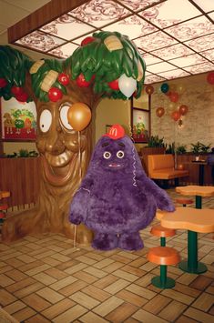 a large purple stuffed animal sitting next to a tree in a room with tables and chairs