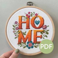 someone is holding up a cross - stitch hoop with the word home in orange letters