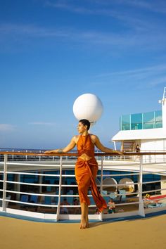 a woman in an orange dress standing on the deck of a cruise ship holding a large white ball over her head