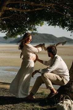 a man and woman sitting under a tree on the beach next to a body of water