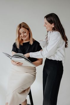 two women are sitting on stools and one is holding a magazine while the other holds a glass of wine