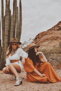 two women sitting next to each other in front of large cactus trees and rocks, one wearing a hat