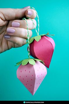 a hand holding two paper strawberries with string attached to the top, on a blue background