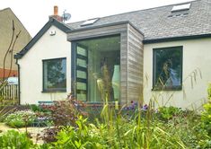 a woman standing in the open window of a small white house surrounded by greenery