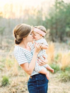 a woman holding a baby in her arms and kissing it's face with trees in the background