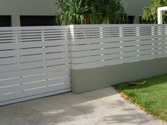 a large white fence next to a house with grass on the ground and palm trees behind it