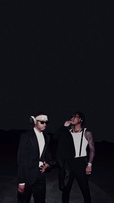 two men standing next to each other in front of a dark sky with one man wearing sunglasses