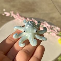 a hand holding a small toy octopus on top of a white plate with pink flowers in the background