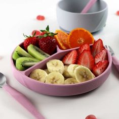 a pink bowl filled with sliced bananas, strawberries and oranges next to two spoons