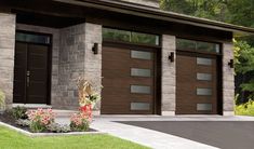 two brown garage doors in front of a brick building with flowers on the lawn and trees behind them