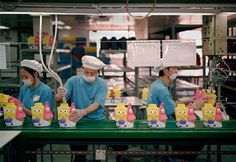 three workers in blue shirts and white hats are working on plastic toy figures that look like spongebob heads