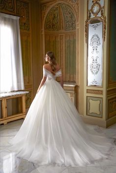 a woman in a white wedding dress is standing near a window and looking off to the side