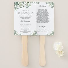 two wooden spatulas with wedding programs on them next to a white flower and greenery