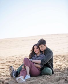 a man and woman sitting in the sand on top of a sandy beach with their arms around each other