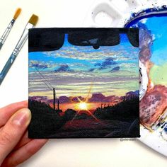 someone is holding up a small painting with the sun setting in the background