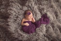 a newborn baby wearing a purple wrap and headband is laying on a gray blanket