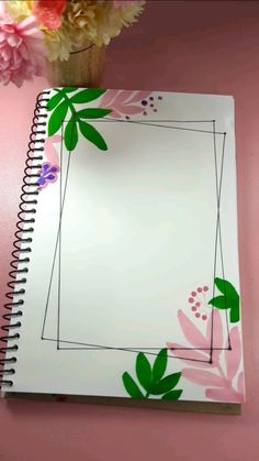 an open notebook with flowers on it next to a pink wall and a vase full of flowers