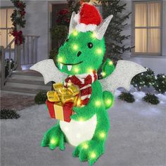 a green dragon holding a present in front of a house with christmas lights on it