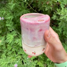 a hand holding up a pink and white cup in front of some green plants with red berries on it