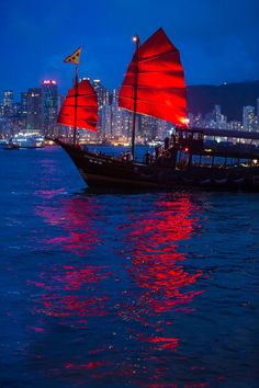 a boat with red sails is in the water near a large city at night time