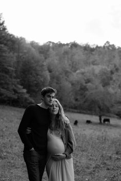 black and white photograph of a pregnant couple standing in a field with cows behind them