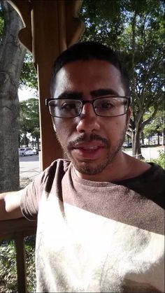 a man wearing glasses and a brown t - shirt is posing for the camera with trees in the background