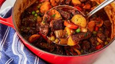 a red pot filled with stew and vegetables on top of a blue towel next to a wooden spoon