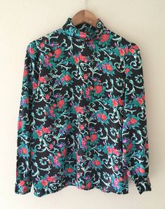 "Vibrant red, pink, and purple flowers, with green foliage on a black background. So pretty!  This vintage, silky blouse is untagged but feels like satin polyester. My guess at the brand is that it's Petite Sophisticates as it is similar in make to the other listings I have from the same lady's estate. (feel free to check my shop for similar listings) There are 5 black, faceted buttons at the back neck closure and 1 at each wrist. It is an approximate size 10 or large - please see measurements below: 22\" underarm to underarm 14.5\" shoulder to shoulder 23\" length front  25.5\" length back 20.5 across bottom 24.5\" sleeve" Pink And Purple Flowers, Silky Blouse, Green Foliage, Pink And Purple, Floral Blouse, Vibrant Red, Purple Flowers, Black Background, Black Backgrounds