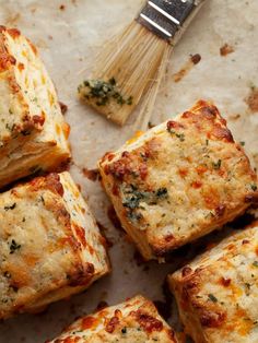 several square pieces of food with cheese and herbs next to a whisk on a piece of parchment paper