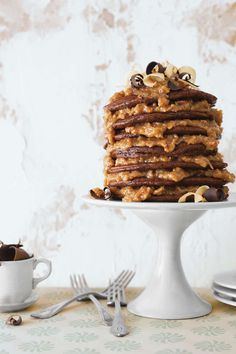 a stack of pancakes with chocolate and nuts on top sitting on a white cake stand