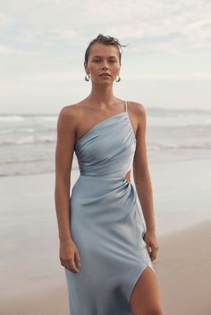 a woman is standing on the beach in a blue one - shoulder dress with thigh high slits