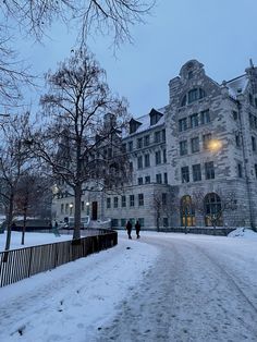 two people walking down a snow covered road in front of a large building with many windows