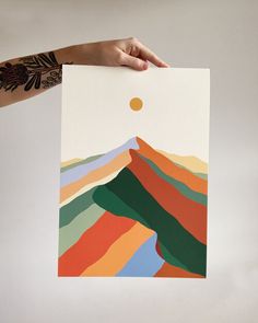 a person holding up a piece of art with mountains in the background and an orange dot above it