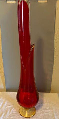 a red glass vase sitting on top of a white tablecloth covered table with a gold plated base