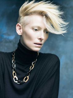 a woman with blonde hair wearing a black turtle neck sweater and gold chain necklaces