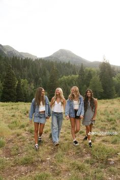 four young women walking through a field with trees in the background and mountains in the distance