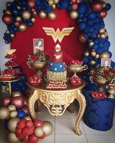 a table topped with lots of balloons and cake next to a wall covered in wonder woman decorations