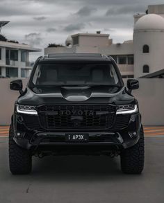 the front end of a black truck parked on top of a parking lot with buildings in the background