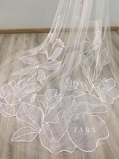 a white veil with flowers on it sitting on top of a wooden floor next to a wall