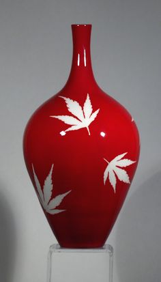 a red vase with white leaves painted on the front and sides, sitting on a clear stand