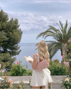 a woman standing in front of some flowers and trees reading a book with the ocean in the background