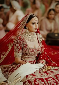 a woman in a red and gold bridal gown sits on the floor with other people behind her