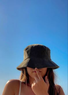 a woman wearing a hat standing on the beach with her finger up to her nose