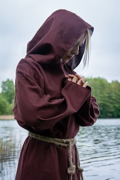 a woman in a hooded robe standing by the water with her hands clasped to her chest