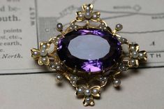 A beautiful  antique Late Victorian to Edwardian 14k yellow gold large  amethyst and seed pearl  brooch.  A lovely design with shamrocks at the four corners and the gold work set with 44 tiny seed pearls.   The brooch measures 1 5/8"  x  1 1/2". There is a large oval faceted amethyst in the center prong set and measures approx. 21mm  x  15mm  x 10mm  (16.7ct).   Set down into the gold are 44 small seed pearls with the size ranging around .75mm to 2.5mm .  The back is fitted with a ball hinge and Elegant Purple Gemstone Brooches, Elegant Amethyst Brooches For Wedding, Purple Gemstone Wedding Brooches, Purple Gemstone Brooches For Weddings, Elegant Purple Brooches For Anniversary, Antique Purple Brooches For Formal Occasions, Elegant Gold Amethyst Brooches, Purple Victorian Wedding Brooches, Gold Work