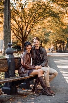 a man and woman are sitting on a bench in the park, posing for a photo