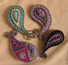 three embroidered brooches are sitting on a cloth covered surface, one is purple and the other is green