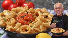 a man holding a plate of food next to a bowl of pasta and tomatoes on a table