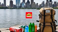 a backpack, water bottles and other items are sitting on the ground next to a body of water with a city in the background